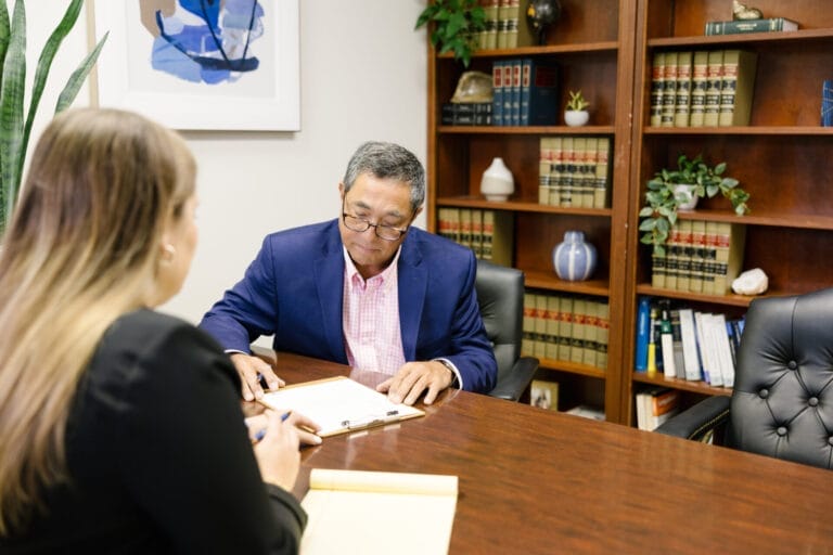A man sits at a desk looking at a clipboard. A woman sits in the foreground across the table from him.